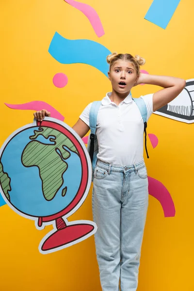 Écolière surprise toucher la tête tout en tenant la maquette globe près du crayon de papier découpé et des éléments abstraits sur jaune — Photo de stock