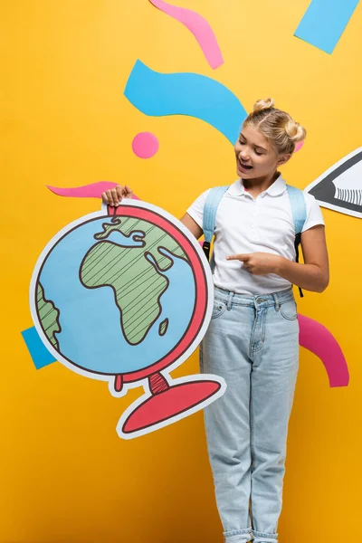 Colegiala emocionada señalando con el dedo en maqueta globo cerca de lápiz de papel cortado y elementos de colores en amarillo - foto de stock