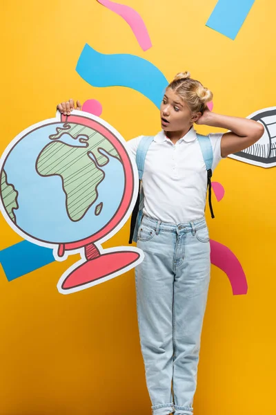 Surprised schoolgirl touching head while holding globe maquette near paper pencil and decorative elements on yellow — Stock Photo