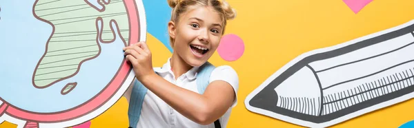 Panoramic shot of excited schoolgirl holding maquette of globe on yellow background with paper elements and pencil — Stock Photo