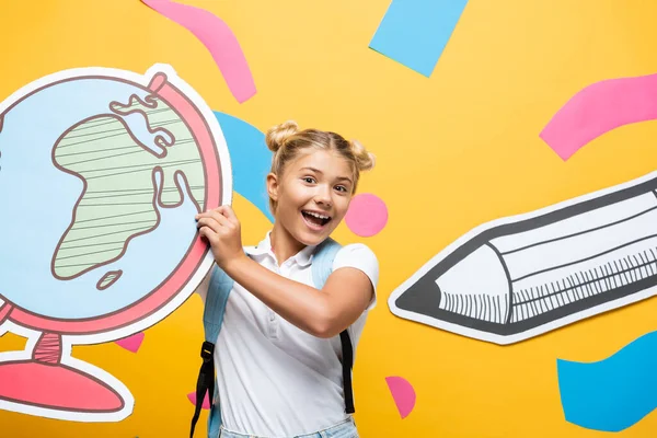 Aufgeregtes Schulkind hält Globus-Maquette in der Nähe abstrakter Elemente und Bleistift auf gelbem Papier — Stockfoto