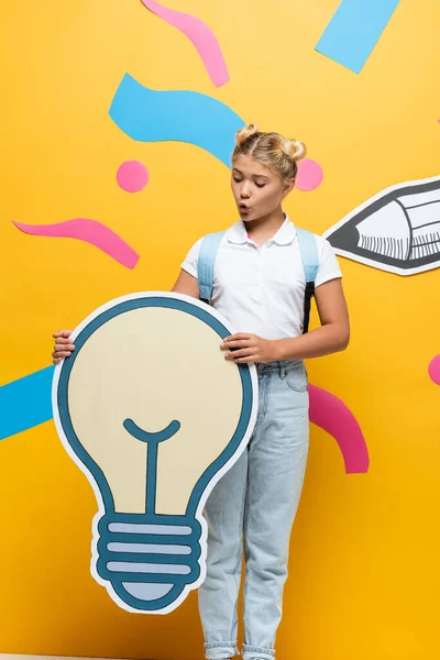 Amazed pupil holding light bubble maquette near multicolored elements and paper pencil on yellow — Stock Photo