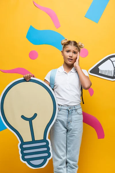 Schockierte Pupille berührt Gesicht, während sie leichte Blasenmaquette auf gelbem Hintergrund mit dekorativen Elementen und Papierstift hält — Stockfoto