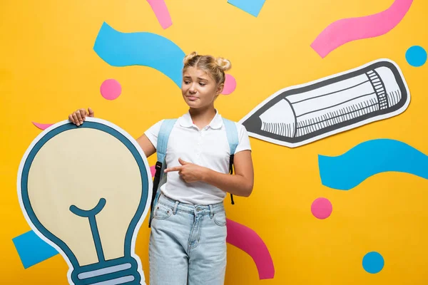 Confused schoolgirl pointing at paper light bulb near paper art on yellow background — Stock Photo