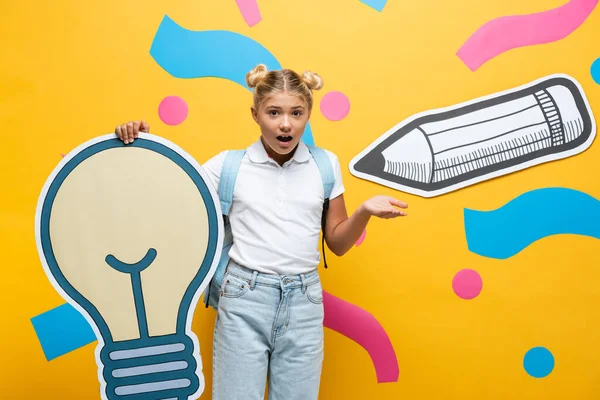 Verwirrtes Schulmädchen zeigt mit der Hand, während sie Papierglühbirnen auf gelbem Hintergrund hält — Stockfoto