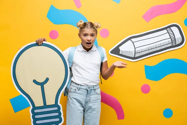 Colegiala confundida apuntando con la mano mientras sostiene la bombilla de papel oso arte de papel sobre fondo amarillo - foto de stock