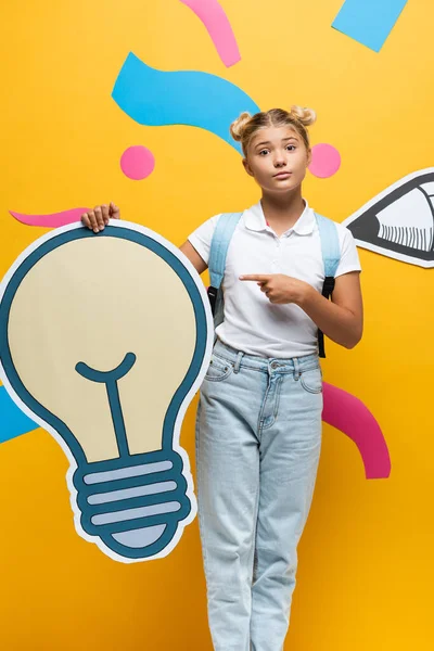 Schoolgirl with backpack pointing with finger at decorative light bulb near paper art on yellow background — Stock Photo