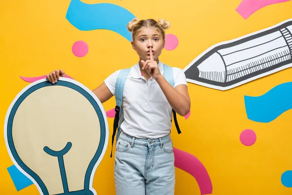 Schoolgirl showing quiet gesture while holding decorative light bulb near paper art on yellow background — Stock Photo