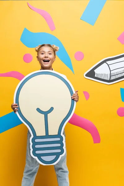 Excited schoolgirl holding paper light bulb beside paper elements on yellow background — Stock Photo