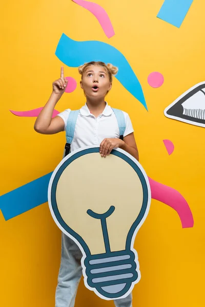 Umfassende Schülerin mit Idee, während sie dekorative Glühbirne in der Nähe von Papierkunst auf gelbem Hintergrund hält — Stockfoto