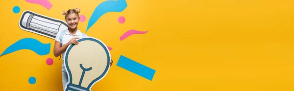 Cultura horizontal de estudante segurando lâmpada decorativa perto de arte de papel no fundo amarelo — Fotografia de Stock