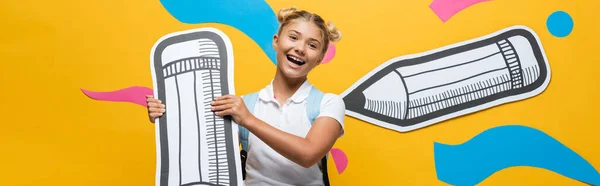 Panoramaaufnahme eines Schulkindes mit Rucksack, der einen dekorativen Bleistift mit Papierkunst auf gelbem Hintergrund hält — Stockfoto
