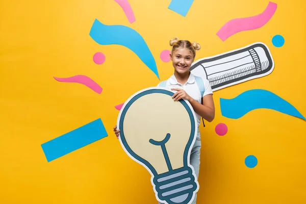 Schulkind blickt in die Kamera, während es eine Glühbirne in der Nähe von Papierkunst auf gelbem Hintergrund hält — Stockfoto