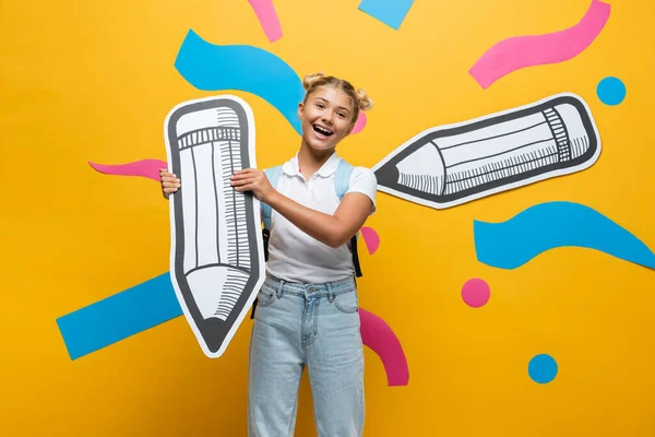 Colegiala con mochila sosteniendo lápiz de papel al lado del arte de papel sobre fondo amarillo - foto de stock