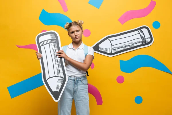 Triste écolière tenant crayon de papier et regardant la caméra sur fond jaune — Photo de stock