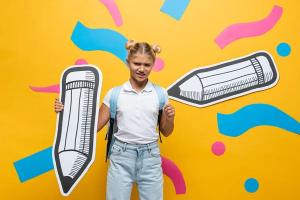 Displeased schoolchild with backpack and paper pencil on yellow background — Stock Photo