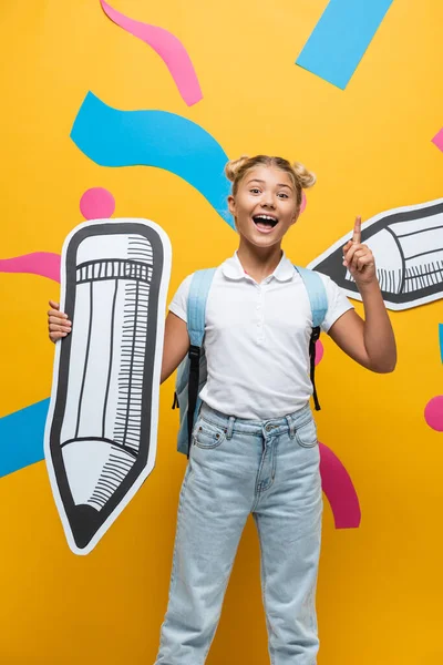 Aufgeregte Schülerin mit Idee, während sie Papierkunst auf gelbem Hintergrund hält — Stockfoto