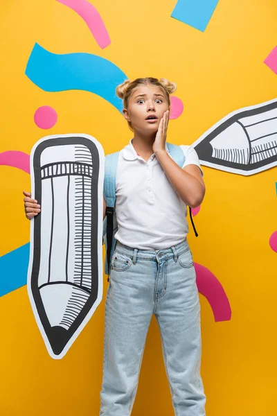 Besorgtes Schulkind mit Rucksack und Bleistift blickt in die Kamera auf gelbem Hintergrund — Stockfoto