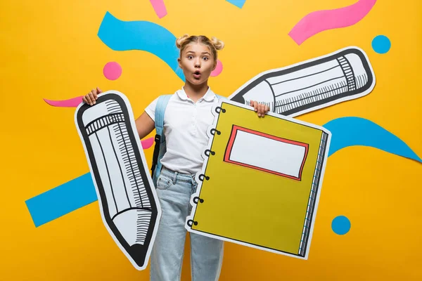 Surprised schoolgirl holding paper pencil and notebook on yellow background — Stock Photo