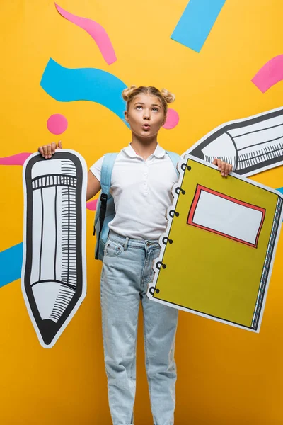 Pensive schoolgirl holding paper pencil and notebook near paper art on yellow background — Stock Photo