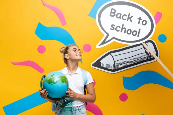 Écolière en lunettes tenant globe près des éléments en papier avec lettrage de retour à l'école sur bulle de parole sur fond jaune — Photo de stock
