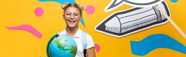 Horizontal image of schoolkid with globe looking at camera near paper craft on yellow background — Stock Photo