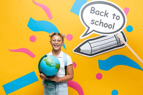 Colegial con globo mirando a la cámara cerca del arte de papel con letras de regreso a la escuela sobre fondo amarillo - foto de stock