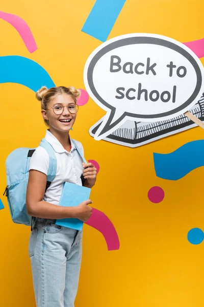 Школа с рюкзаком и книгой, стоящей рядом с речевым пузырём со школьными надписями и бумажными рисунками на жёлтом фоне — стоковое фото