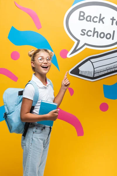 Aufgeregtes Schulmädchen mit Buch zeigt auf Sprechblasenwitz zurück zu Schulbuchstaben und Papierkunst auf gelbem Hintergrund — Stockfoto
