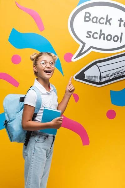 Colegiala emocionada con el libro apuntando al ingenio de la burbuja del habla de nuevo a las letras de la escuela y el arte del papel en el fondo amarillo - foto de stock