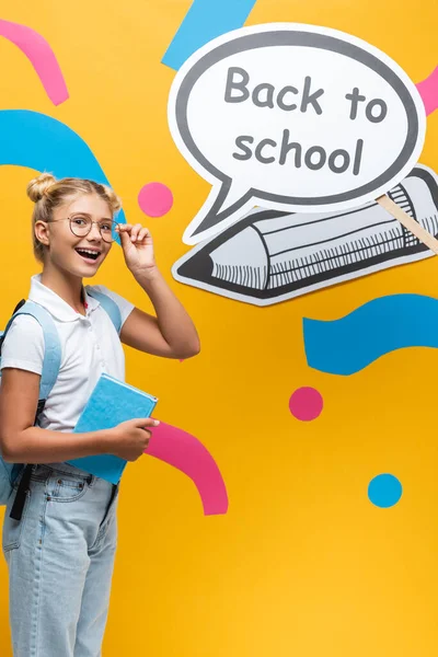 Школьник с рюкзаком и очками стоит рядом с речевым пузырём со школьными надписями и бумажными изделиями на жёлтом фоне — стоковое фото