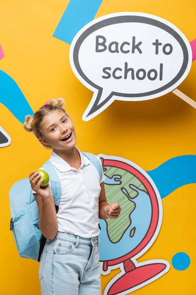 Schoolchild holding apple near speech bubble with back to school lettering and paper elements on yellow background — Stock Photo
