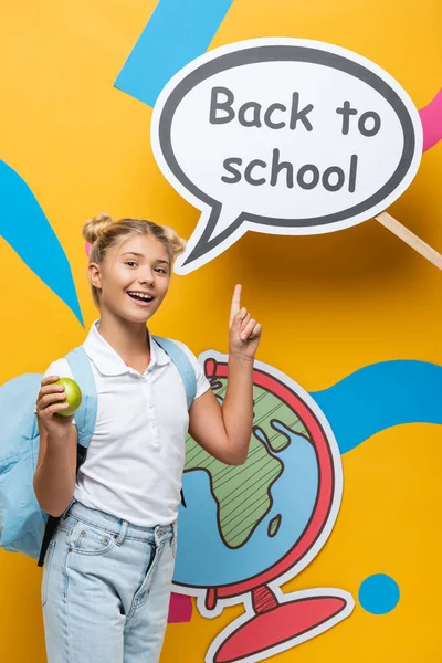 Школьник с рюкзаком и яблоком указывает на бумажный пузырь речи со школьными надписями на желтом фоне — стоковое фото