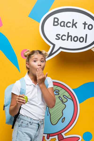 Colegial sorprendido sosteniendo manzana y cubriendo la boca cerca de la burbuja del habla con letras de regreso a la escuela sobre fondo amarillo - foto de stock