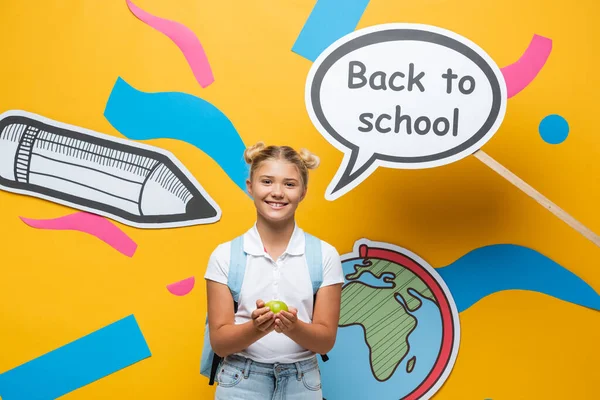 Шкільний малюк тримає яблуко біля паперового мистецтва та мовної бульбашки зі спиною до шкільного приладдя на жовтому фоні — стокове фото