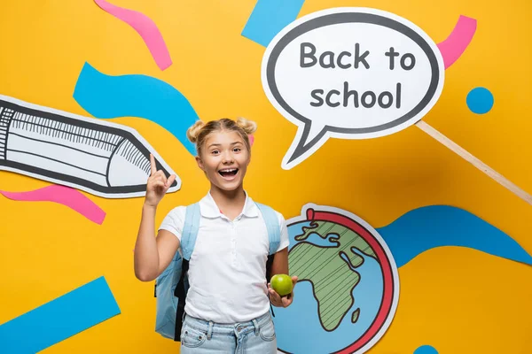 Colegiala con manzana tener idea cerca de vuelta a la escuela letras en la burbuja del habla y el arte del papel sobre fondo amarillo - foto de stock