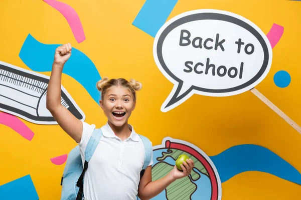 Écolière excitée avec pomme montrant geste ouais près de bulle de parole avec retour à l'école lettrage et art du papier sur fond jaune — Photo de stock