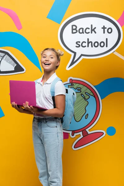 Colegiala sosteniendo portátil cerca de la burbuja del habla con letras de vuelta a la escuela y el arte del papel sobre fondo amarillo - foto de stock