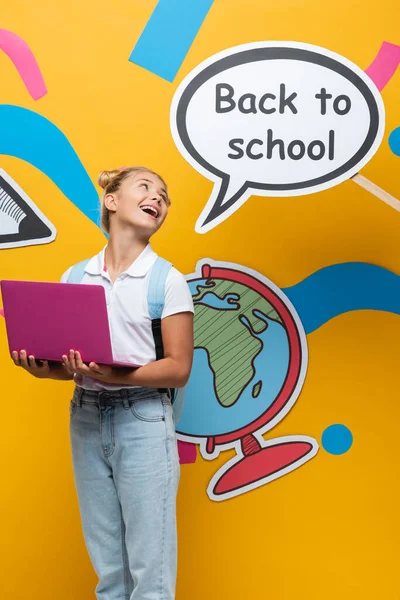 Scolaretta con computer portatile che guarda la bolla vocale con lettere di ritorno a scuola e arte cartacea su sfondo giallo — Foto stock