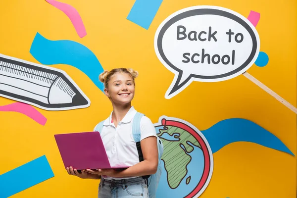 Schulmädchen mit Laptop und Rucksack blickt in die Kamera neben Papierkunst und Sprechblase mit Schulbuchstaben auf gelbem Hintergrund — Stockfoto