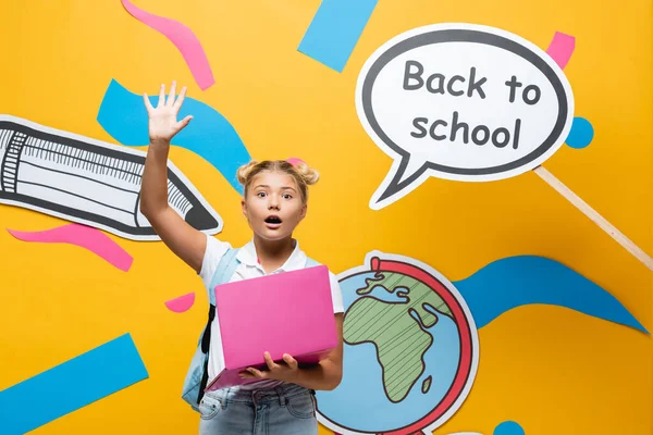 Écolière excitée avec ordinateur portable agitant à la caméra près de bulle de parole avec retour à l'école lettrage et art du papier sur fond jaune — Photo de stock