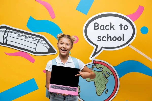 Colegial apuntando a la computadora portátil con pantalla en blanco cerca de la burbuja del habla con letras de vuelta a la escuela y el arte del papel sobre fondo amarillo - foto de stock