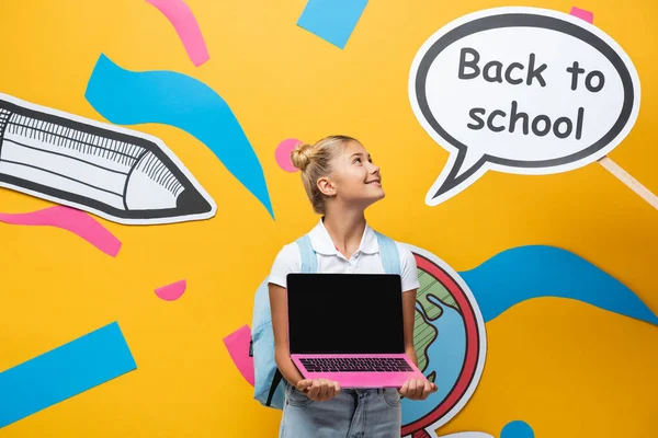 Schoolgirl with backpack and laptop looking at speech bubble with back to school lettering and paper art on yellow background — Stock Photo