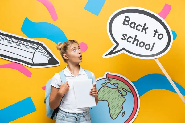 Écolier excité avec sac à dos et tablette numérique regardant la bulle de la parole avec le lettrage de retour à l'école et l'art du papier sur fond jaune — Photo de stock