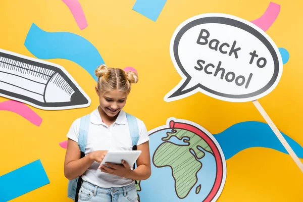 Schoolkid usando tablet digital perto da bolha de fala com letras de volta para a escola e arte de papel no fundo amarelo — Fotografia de Stock