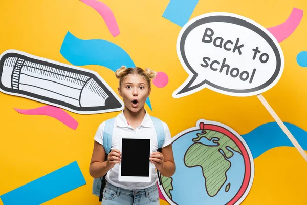 Шокированная школьница держит цифровой планшет рядом с бумажным искусством и мыльным пузырем речи на желтом фоне — стоковое фото
