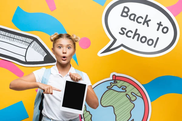 Surprised child pointing with finger at digital tablet near speech bubble and paper art on yellow background — Stock Photo