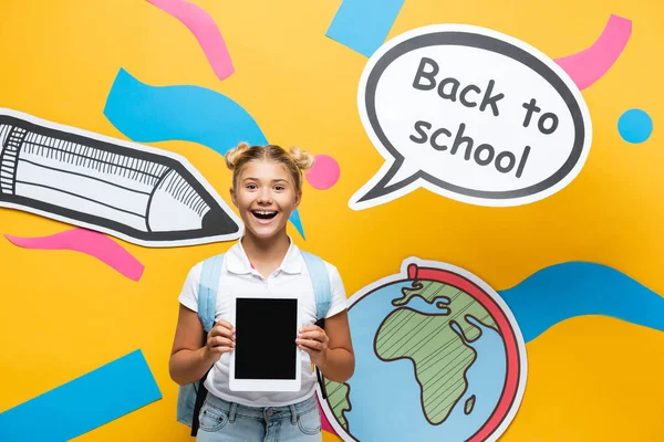 Colegial sosteniendo tableta digital con pantalla en blanco cerca de papel artesanal y burbuja del habla sobre fondo amarillo - foto de stock