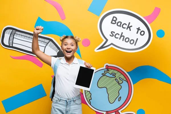 Schoolgirl holding digital tablet and showing yeah gesture near speech bubble and paper art on yellow background — Stock Photo