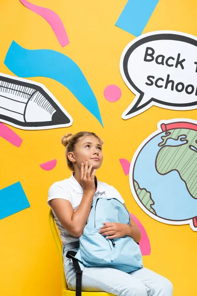 Colegiala soñadora sosteniendo la mochila en la silla cerca de la burbuja del habla y el arte del papel sobre fondo amarillo - foto de stock